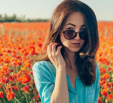 Beautiful brunette young woman in sunglasses in spring red poppies flowers meadow, looking at camera.