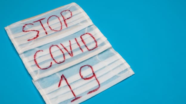 Typical 3-ply white surgical mask with rubber earhooks to cover mouth and nose with English block letters Stop Covid-19 on a blue background. Dangerous virus and quarantine concept