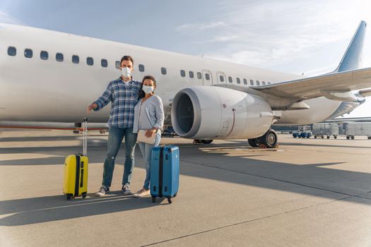 Full length photo of adult couple wearing protective masks and standing near the plane and going to travel. Coronavirus concept