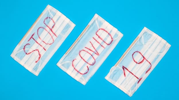 Typical 3-ply white surgical mask with rubber earhooks to cover mouth and nose with English block letters Stop Covid-19 on a blue background. Dangerous virus and quarantine concept.