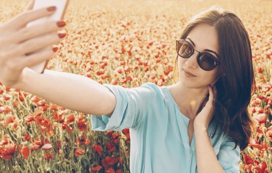 Pretty stylish young woman taking selfie with smartphone in poppies flowers meadow in spring outdoor.