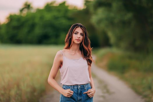 Portrait of beautiful sexy young woman with long brown hair in pink tank top and denim shorts posing outdoors at summer sunset, sensual, serious, blurry nature background