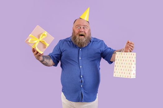 Amazed middle aged bearded man with overweight in party hat holds gift box and paper bag on purple background in studio