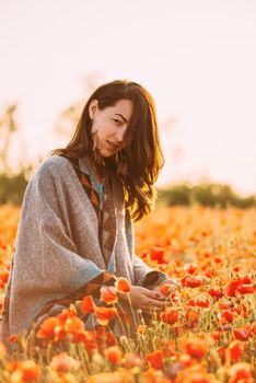 Beautiful young woman walking in red poppies flowers meadow in spring outdoor, looking at camera.