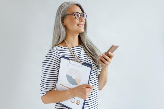 Cheerful mature Asian woman with loose hair holds diagrams and mobile phone standing on light grey background in studio