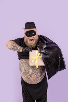 Excited mature bearded man with overweight and tattoos on large belly in Zorro costume hides gift box behind black cape on purple background in studio