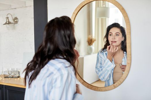 Young brunette looking at mirror and smearing lipstick on lips while applying makeup in morning at home