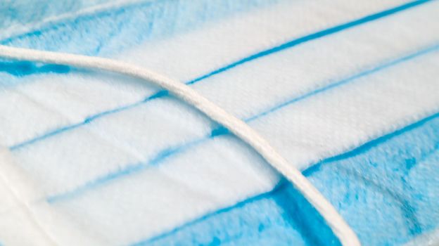 One 3-ply disposable surgical face mask close-up with rubber pads covering mouth and nose against blue background. The concept of protection from bacteria, healthcare and medicine. COVID-19