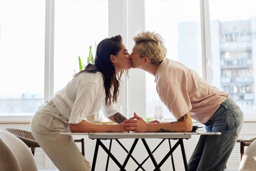 Side view of young women in casual clothes holding hands and kissing each other over table in morning at home