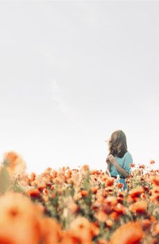 Romantic young woman walking in red poppies meadow and sniffing a flower in spring outdoor. Copy-space in upper part of image.