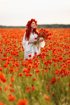 Portrait of beautiful redhead woman in wreath on green field with bouquet of poppies in summer sunset
