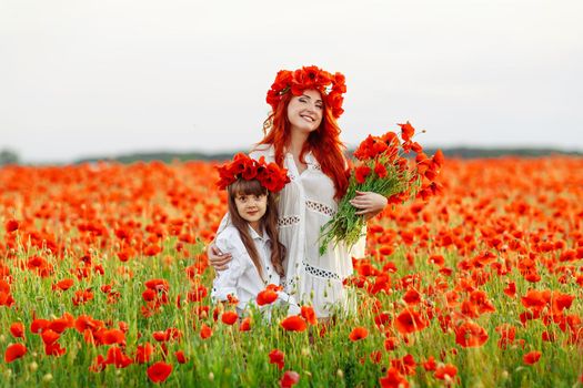 Little girl with redhead mother in white dresses and wreathes poses with bouquet of poppies on poppy field at warm summer sunset