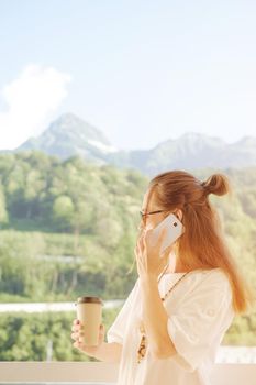Young woman standing on balcony with cup of coffee and talking on phone, mountains in the background.