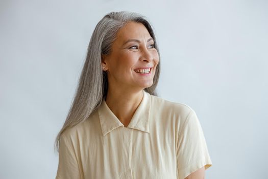 Joyful long haired middle aged lady wearing beige blouse stands on light background in studio. Mature beauty lifestyle