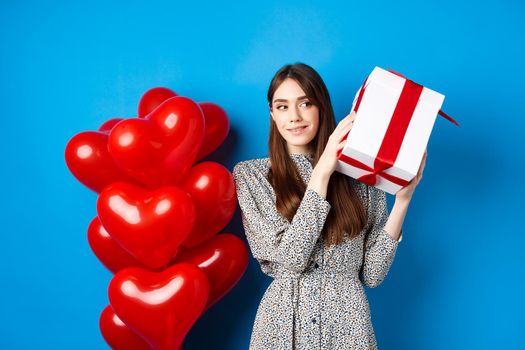 Valentines day. Beautiful woman shaking gift box to guess what inside, celebrating lovers holiday, standing near red hearts, blue background.