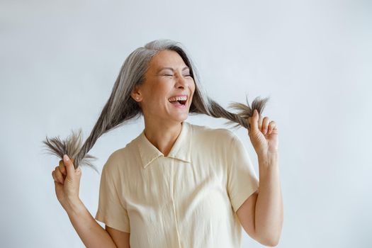 Joyful Asian lady in blouse holds long hoary hair laughing on light grey background in studio. Mature beauty lifestyle