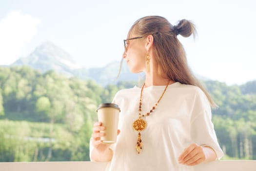 Beautiful young woman with cup of coffee enjoying view of mountains on resort outdoor.