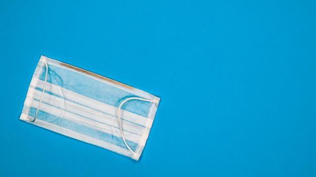 One 3-ply disposable surgical face mask with rubber pads to cover mouth and nose against blue background. The concept of protection from bacteria, healthcare and medicine. COVID-19. copy space