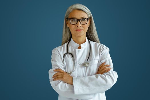 Mature female doctor in white robe with glasses and stethoscope stands on blue background in studio. Professional medical staff