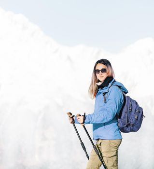 Backpacker explorer young woman with trekking poles walking in front of mountains in winter.