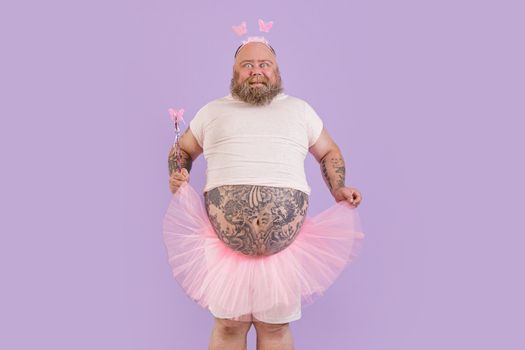 Excited obese mature man with large bare abdomen wearing fairy suit holds magic stick and chiffon skirt on purple background in studio