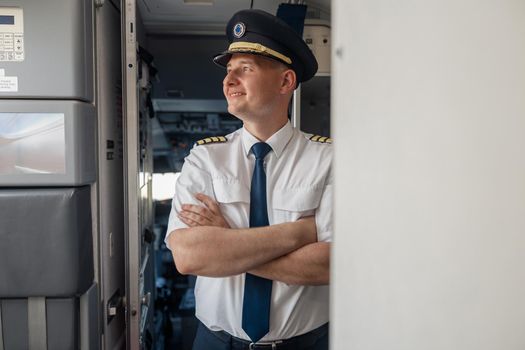 Dreamy male pilot in uniform and hat keeping arms crossed and smiling away while posing, standing inside of the airplane. Transportation, aircrew concept