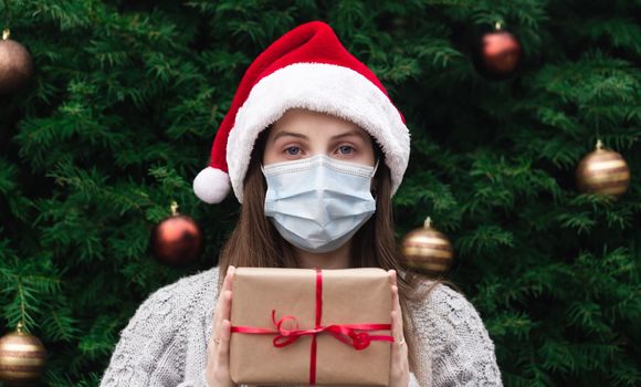 Christmas mask congratulations. Portrait woman wearing santa hat and sweater in medical mask, giving gift present box with red ribbon, christmas tree bokeh on background