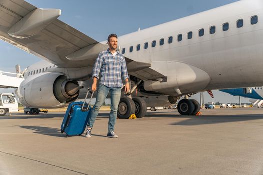 Happy adult man walking with travel bag near big white airplane. Trip concept