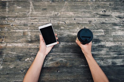 Point of view of female hands holding smartphone and takeaway cup of coffee on wooden background, mock-up.