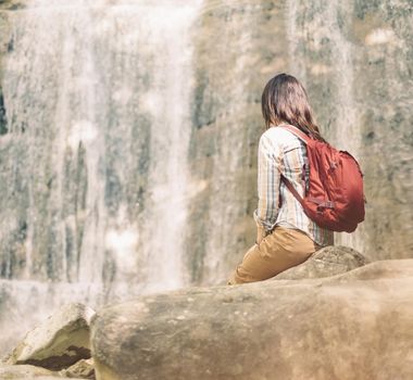 Unrecognizable traveler backpacker young woman sitting on stone and looking at waterfall on sunny day outdoor.
