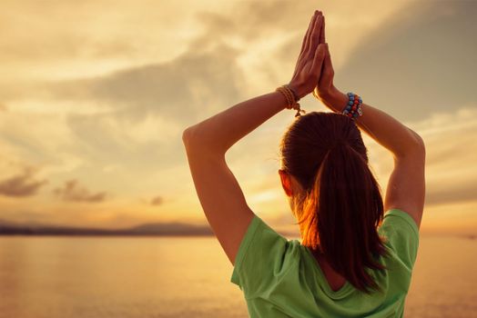 Unrecognizable young woman practicing yoga with praying gesture in front of sea at sunset outdoor.