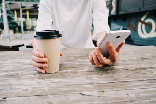 Unrecognizable young woman sitting with cup of coffee and mobile phone at wooden table in cafe in city street.