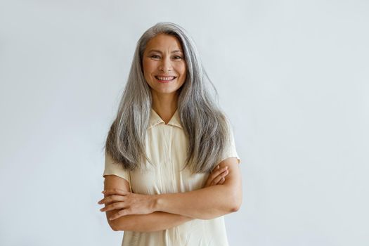 Portrait of smiling Asian woman with long hoary hair and crossed arms standing on light grey background in studio. Mature beauty lifestyle