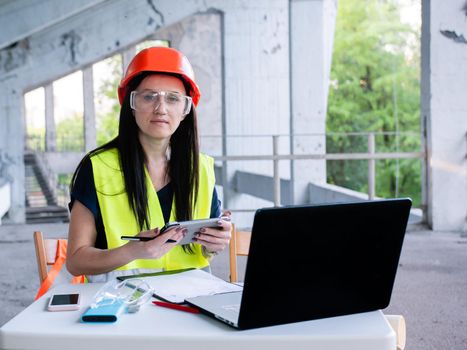 A female builder worker in an orange helmet sits on a construction site with a computer. The foreman checks the home renovation process.