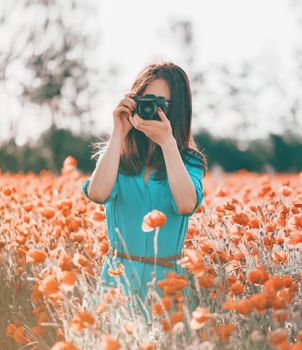 Brunette young woman taking a photo with a camera in poppy flower meadow in summer outdoor.