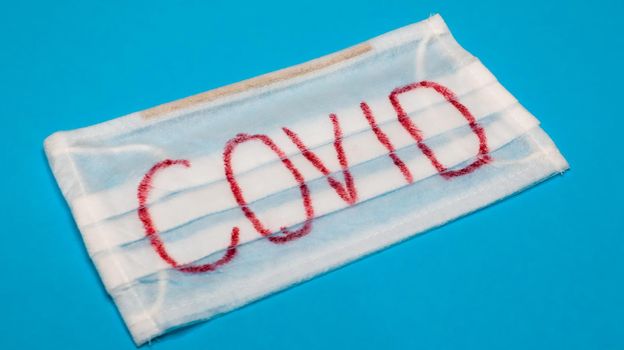 Typical white 3-ply surgical mask with rubber earhooks to cover mouth and nose with English block letters Covid on a blue background. Dangerous virus and quarantine concept