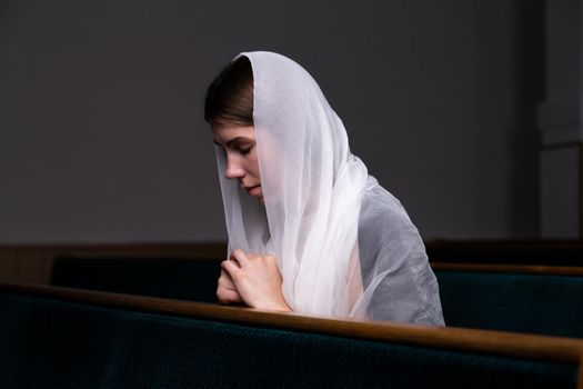 A young modest girl with a handkerchief on her head is sitting in church and praying. The concept of religion, prayer, worship.