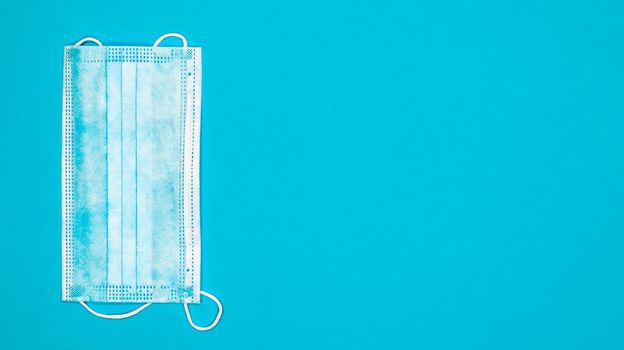 One 3-ply disposable surgical face mask with rubber pads covering the mouth and nose against a blue background. The concept of protection from bacteria, healthcare and medicine. COVID-19. copy space.