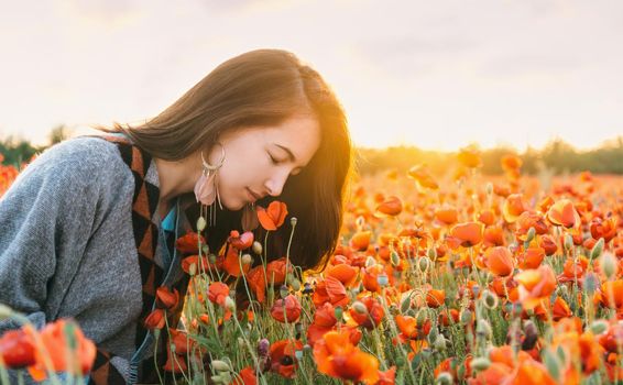 Romantic beautiful brunette young woman smelling a red poppy in flower meadow in summer sunset.
