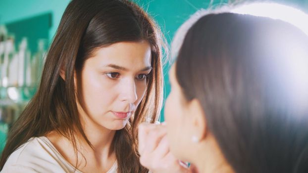 Professional stylist female doing make-up for young woman in front of mirror in beauty salon, close up shot