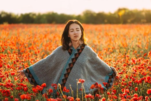 Boho stylish dreamy beautiful young woman relaxing with closed eyes in red poppy flower meadow in summer.