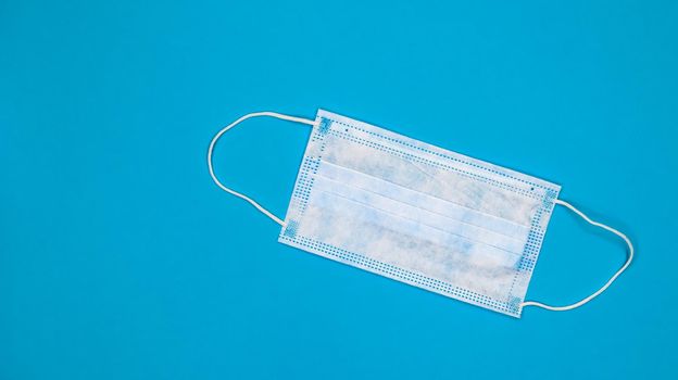 One 3-ply disposable surgical face mask with rubber grips covering mouth and nose against blue background. Protection from bacteria, healthcare and medicine concept. COVID-19. A dangerous virus
