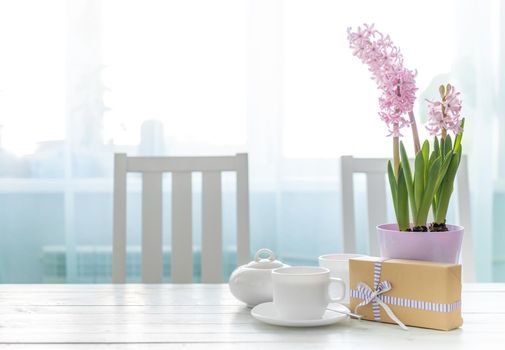 Present gift box with white cups of tea and pink flowers on the white table near window. Lovely spring holiday mood