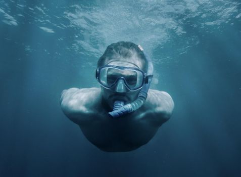 Handsome sporty young man with snorkel and mask swimming underwater, looking at camera.