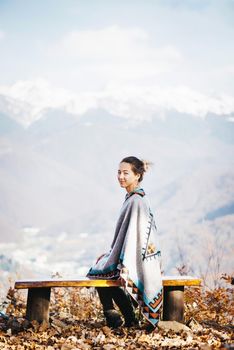 Beautiful young woman sitting on bench in front of snowy mountain ridge, looking at camera.