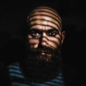 Close-up portrait of middle aged brutal bearded man with expressive eyes and striped shadows on his face with black background