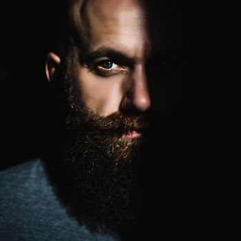 Close-up portrait of middle aged brutal bearded man with expressive eyes and shadow on half of his face with black background