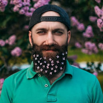 A bearded man with a decorated beard for the Spring holiday. Lilac flowers in the beard.