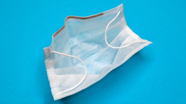 One 3-ply disposable surgical face mask with rubber grips covering mouth and nose against blue background. Protection from bacteria, healthcare and medicine concept. COVID-19. A dangerous virus