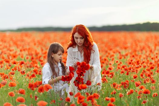 Little happy girl with redhead mother in white dresses makes wreath on poppy field at warm summer sunset